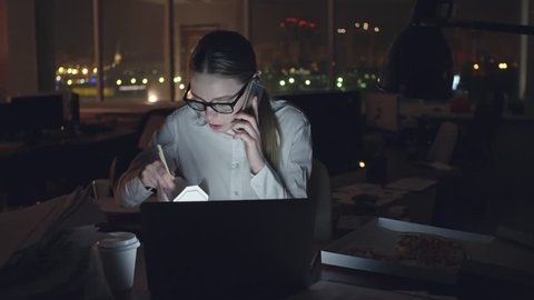 Young businesswoman sitting at desk in dark office, talking on mobile phone and eating noodles from box with sticks, open laptop in front of her, medium shot on Sony NEX700 + Odyssey 7Q