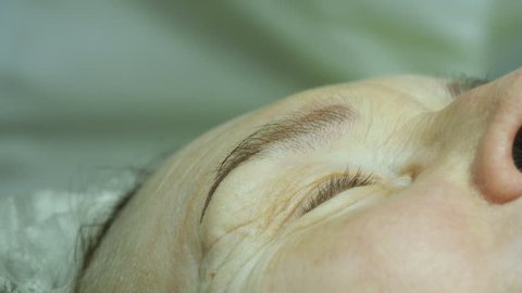 Hands of beautician are preparing the skin for the procedure or cleaning after the procedure. Old woman lies and getting eyelash makeup at beauty salon. Applying Permanent Make up on eyelash.