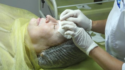 Hands of beautician are preparing the skin for the procedure or cleaning after the procedure. Old woman lies and getting eyelash makeup at beauty salon. Applying Permanent Make up on eyelash.