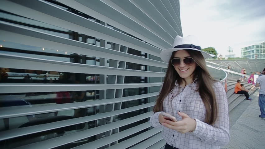Woman is Walking on Street in Sunny Summer Day and Talking on Phone. There Are Lots of People Around Her Who Enjoys Spending Free Time Outside. She Passes by Construction Made From Glass and Metal.