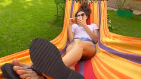 Beautiful caucasian brunette girl swinging in bright hammock and using her smartphone. 4K video