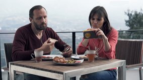 Couple during breakfast on terrace, man listening to music and woman watching movie on smartphone
