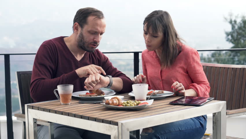 Young couple talking and using smartwatch during breakfast on terrace
