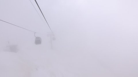Aerial lift pylon and gondolas appear out of heavy fog. POV look out cable car move downward through milklike opaque air, mountains landscape, ski resort elevation system, cloudy weather