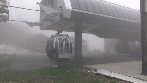 ROZA KHUTOR, RUSSIA - APRIL 29, 2016: Gondola lift descend from cloudy air, pass by pylon and enter station building. Camera turn and follow cabin position. Dull weather at high altitude