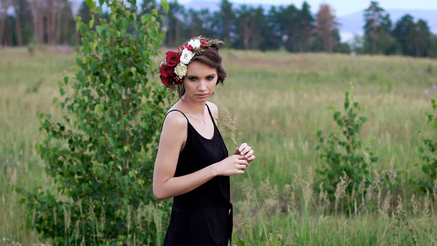 Beautiful girl in the black short dress with the wreath on her head on the green field.