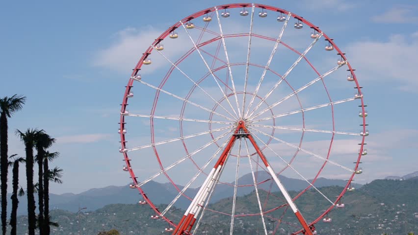 Ferris wheel rotates against the backdrop of palm trees and green hills. Close-up. Batumi, Georgia.

