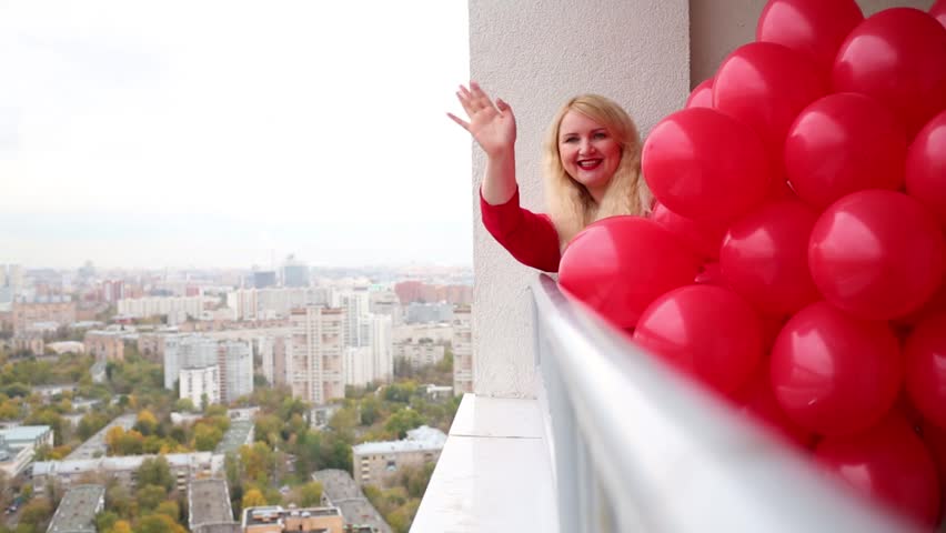 Young woman in red dress is shaking hand and holding balloons on the balcony.