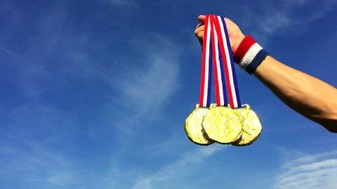 Hand of American athlete holding gold medals hanging from USA colors red, white, and blue ribbons against a blue sky background