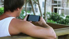 Young man with black phone sits in summer on street, in park, outdoor. Playing on mobile phone, looking at screen. HD, Size: 1080p (1920x1080), Sound: No 