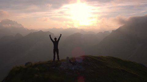 Aerial - Flyover silhouette of a man standing on top of the mountain. Man raising arms victoriously after climbing the mountain