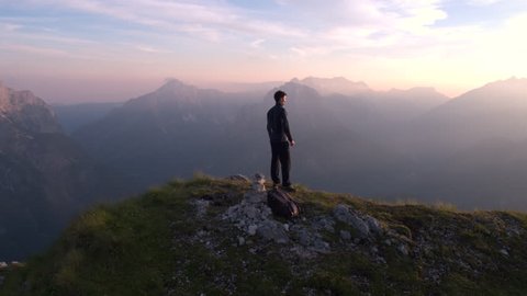Aerial - Flying from front to behind the man standing on top of the mountain watching beautiful sunset over the peaks