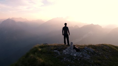 Aerial - Flying around young man standing victoriously on top of the mountain at sunset