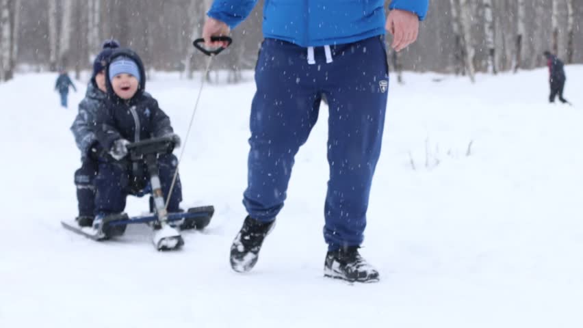 Male hand drags two happy little boys in sled near other boy during snowfall in park
