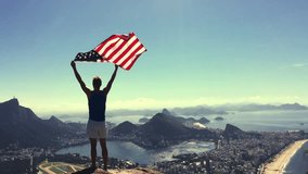 Man stands holding an American flag waving in slow motion at a bright overlook of the city skyline of Rio de Janeiro, Brazil
