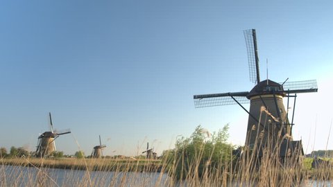 CLOSE UP: View of stunning traditional Dutch windmills from other side of river bank at famous tourist spot Kinderdijk, Netherlands. Beautiful mills on vast plain grassy field near big water stream