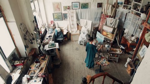 Wide view of a female artist wearing a smock standing in her studio painting a canvas on an easel surrounded by other covered canvasses and art supplies.