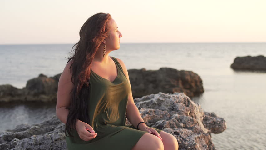 Young attractive woman sitting and relaxing on stones near sea. Middle shot Royalty-Free Stock Footage #18494107