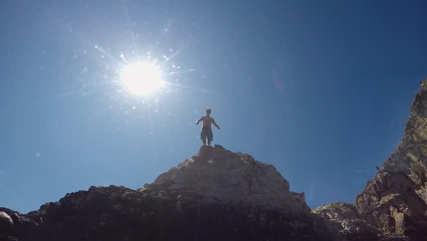 SLOW MOTION UNDERWATER: Happy young man with hands raised jumping into water off a rocky wall at sunny seaside. Fearless guy on fun summer vacation diving into refreshing sea off extremely high cliff