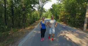 Drone clip of smiling sportwoman and sportsman running along countryside road in sunny forest