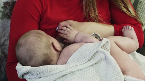 Young mother in red shirt breastfeeding little crying baby on sofa. Mother care. Love
