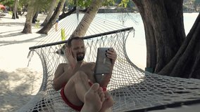 Happy man talking on tablet computer on hammock on beach, super slow motion 120fps
