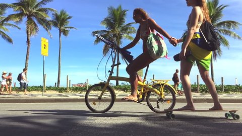 RIO DE JANEIRO - MARCH 20, 2016: A Brazilian woman on bicycle tows a friend on skateboard in slow motion on the beachfront Avenida Vieira Souto road on a car-free weekend afternoon at Ipanema Beach.