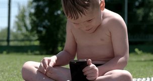 Little Boy With Smartphone or Tablet Sitting in the Garden on Green Grass. Play, Game, Mobile Applications, Children, Yard, Court, Courtyard, Outdoor, Park.