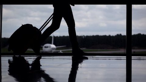 Female passenger legs silhouetted, girl walk with trolley case at airport terminal against glass wall window. Parked airliner seen outside, cloudy sky. Departure gate slow motion shot