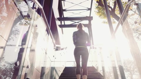 Runner Woman Jogging up Stairs, Lens Flare. SLOW MOTION 120 fps Steadicam STABILIZED shot. Athletic Sportswoman Running Up the Modern Sunny Glass Stairs. Healthy Lifestyle and Wellness. Achievement.