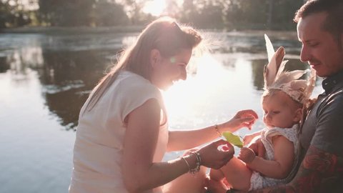Young family with Baby Girl Enjoying Sunny evening in the Park. Slow Motion 120 fps, 4K. Parents relaxing with their toddler daughter near lake. Happy childhood and Parenthood. Love and Happiness