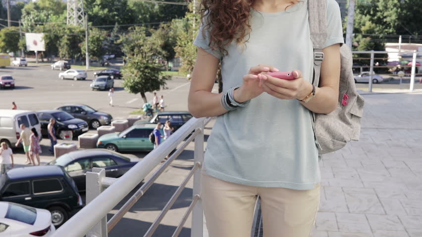 Slender young girl uses a pink mobile phone on city background in slow motion. Woman in a T-shirt with a backpack in the city in the summer.