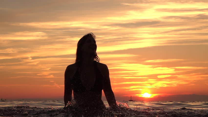 SLOW MOTION, CLOSE UP: Young playful cheerful woman standing in shallow ocean and splashing sea water around with her hands. Happy female sprinkling ocean water at stunning golden sunset