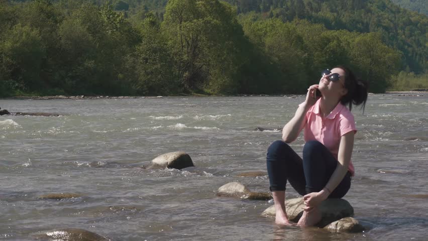 Young attractive brunette girl in sunglasses emotionally talking by phone and smiling. Mountain river background
