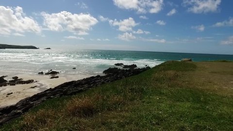A moving pan along a headland in Newquay with view across Fistral Bay beach, tide and surf with distant surfers in Summer.
