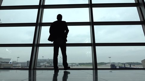 Man come and stand at full height, gaze out airport terminal window, silhouette view. Empty aeroport field outside, hipster person look out while waiting for boarding gate open, VKO lounge hall