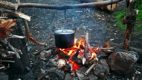 Touristic kettle with boiling water and hot steam over burning flame of campfire among firewood and stones. Full-hd video