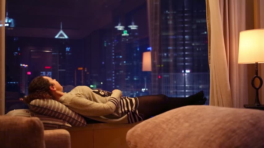 Woman lies on windowsill at night and look at skyscrapers in city