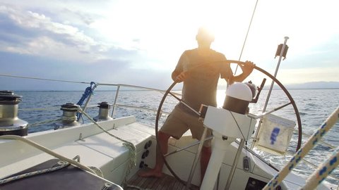 Mature adult man is steering his big sailboat while navigating thru the ocean on a sunny day on his lonely circumnavigation. nice sunset in the back