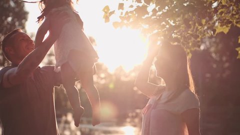 Young family with Baby Girl Enjoying Sunny evening in the Park. Slow Motion 120 fps, 4K. Parents playing with their toddler daughter near lake. Happy childhood and Parenthood. Love and Happiness 