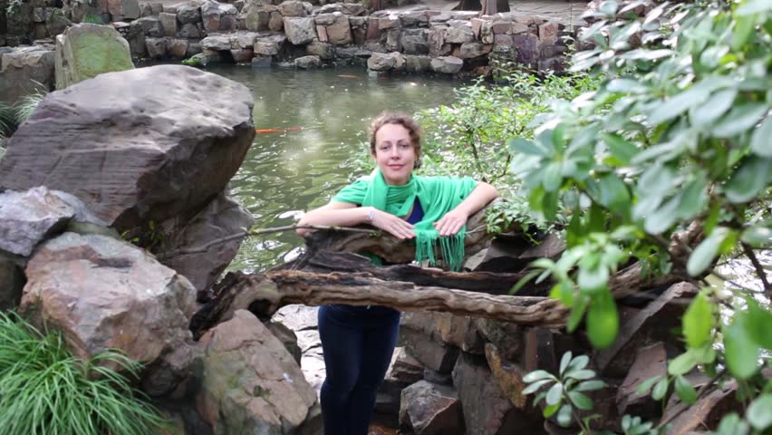 Beautiful woman poses near big stones, pure pond in garden