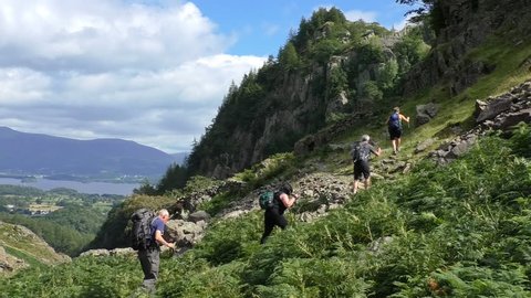 Group of four senior hikers with backpacks and walking trekking poles are climbing mountain slope in Lake District, UK.Beautiful scenery with Derwent Water in the background