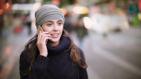 Attractive and cheerful young woman using a smartphone in a crowded street. She is talking on the phone, walking and laughing. Winter time, sunset. 
