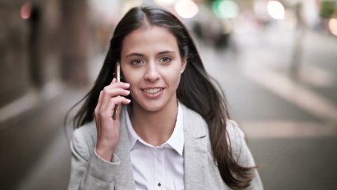 Attractive and cheerful businesswoman using smartphone in a crowded street. She is talking on the phone and laughing while walking. City at sunset.