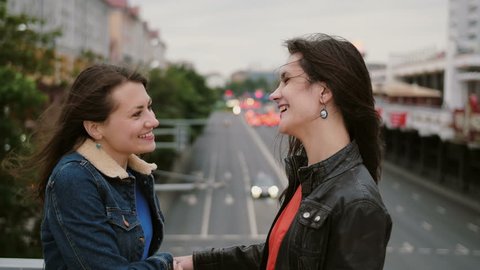 Two beautiful girls best friends met at the city bridge, hugging, kissing, talking, smiling, laughing. 4K