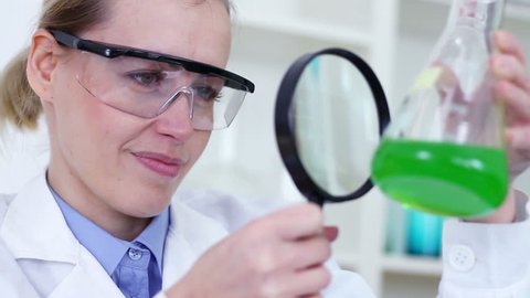 Female scientist with magnifying glass looking at erlenmeyer flask with green chemicals