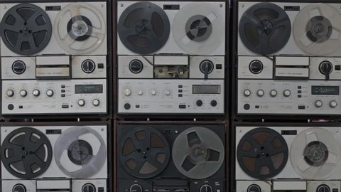 Many vintage magnetic reel to reel tape recorders playing all in the same time, synchronized, with spinning reels. Front view.