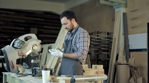 Carpenter with beard makes something on his smart phone in workshop.