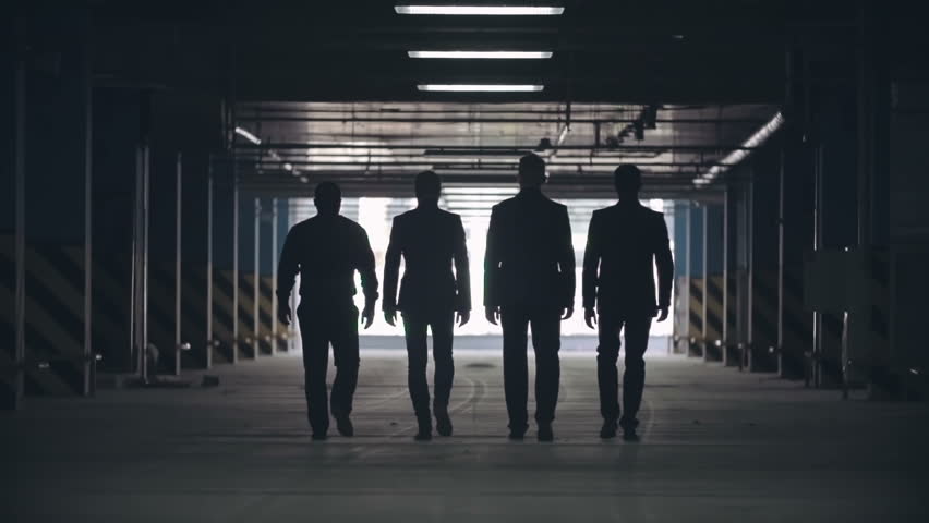Slow motion locked-down rear view of silhouettes of four men in black suits confidently walking away from camera towards exit of parking lot. Royalty-Free Stock Footage #18874913