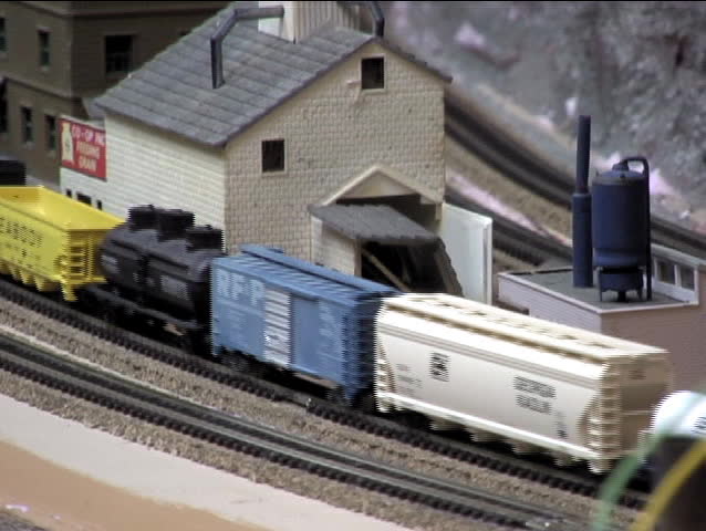 A line of slow model train cars is passed by a faster train after a few seconds.